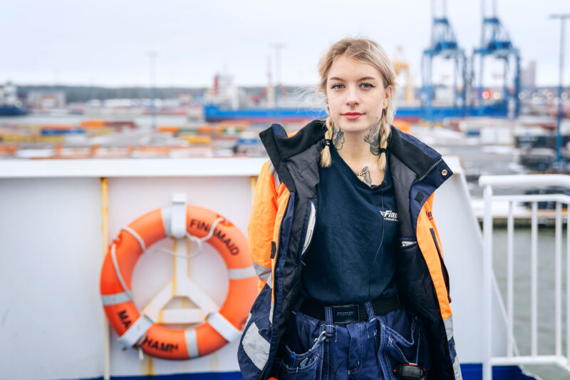 Jemina Lindqvist seisoo laivan kannella ja katsoo suoraan kameraan, taustalla näkyy sataman kontteja ja nostureita.